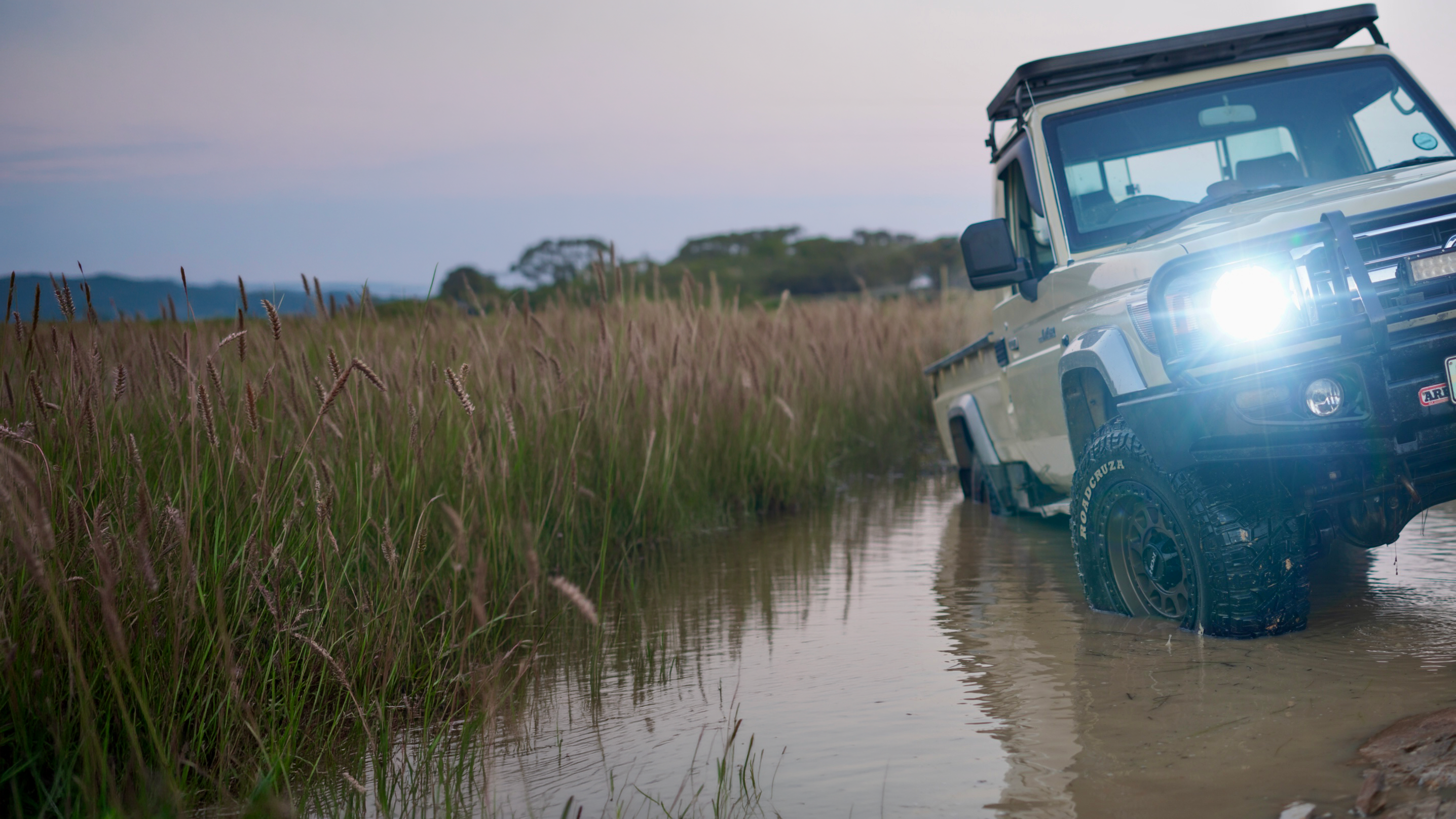 Off-road vehicle driving through a shallow water crossing with tall grasses on either side.